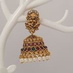 Traditional Gold jhumka earrings with idol design, red and green gemstones and pearl droppings on a white tree branch stand.