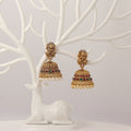 Traditional Gold jhumka earrings with idol design, red and green gemstones and pearl droppings on a white tree branch stand.
