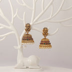 Traditional Gold jhumka earrings with idol design, red and green gemstones and pearl droppings on a white tree branch stand.