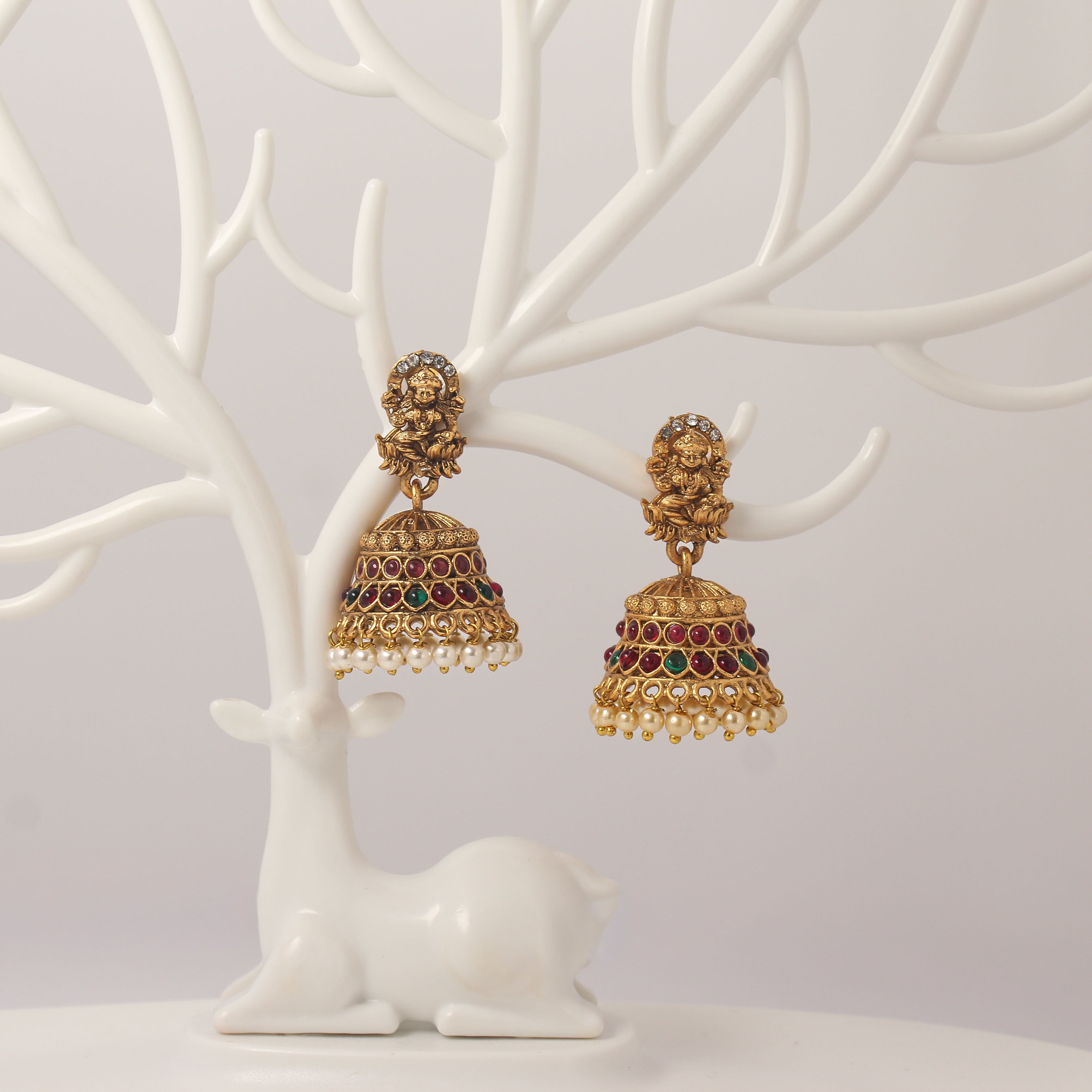 Traditional Gold jhumka earrings with idol design, red and green gemstones and pearl droppings on a white tree branch stand.