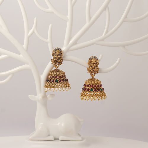 Traditional Gold jhumka earrings with idol design, red and green gemstones and pearl droppings on a white tree branch stand.