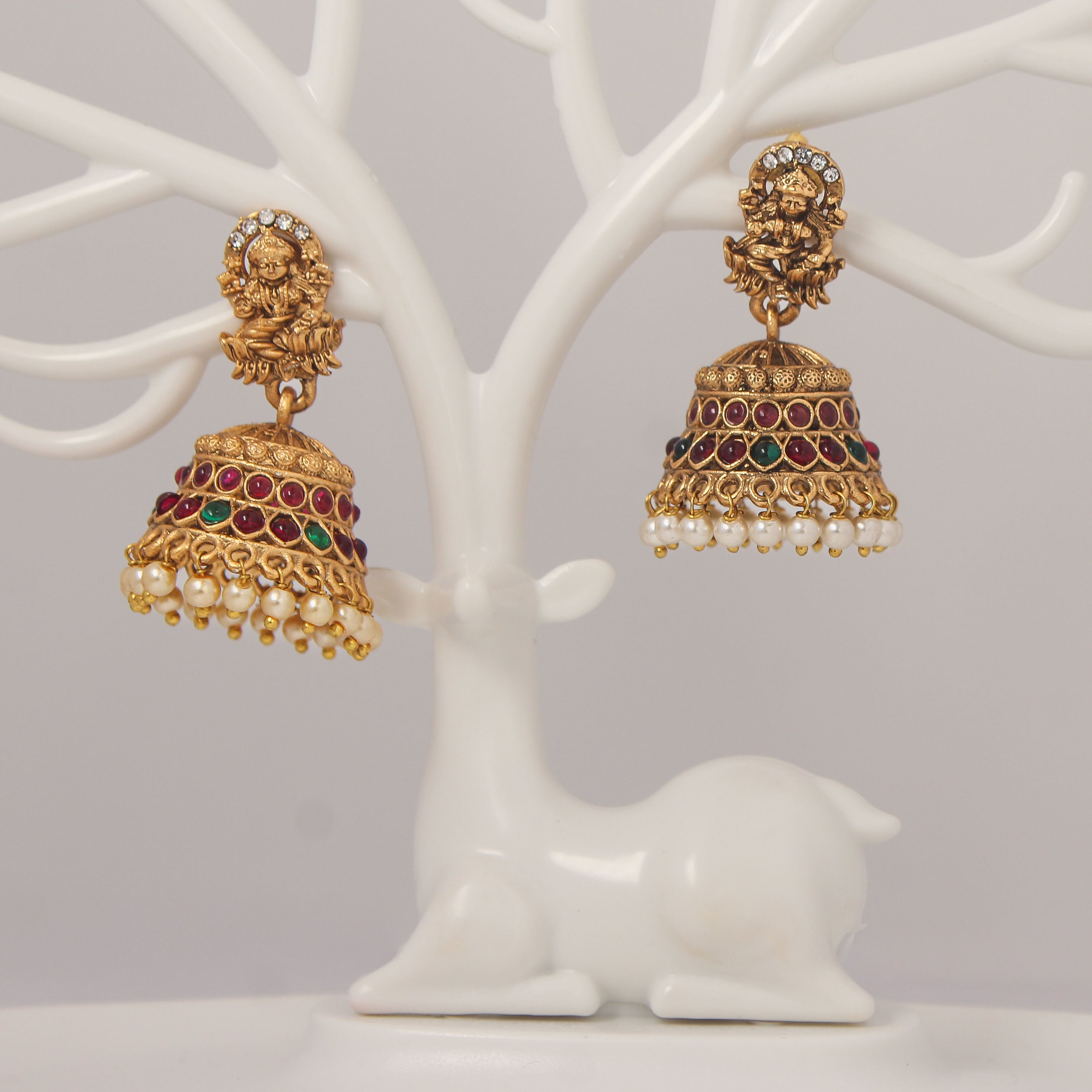 Traditional Gold jhumka earrings with idol design, red and green gemstones and pearl droppings on a white tree branch stand.