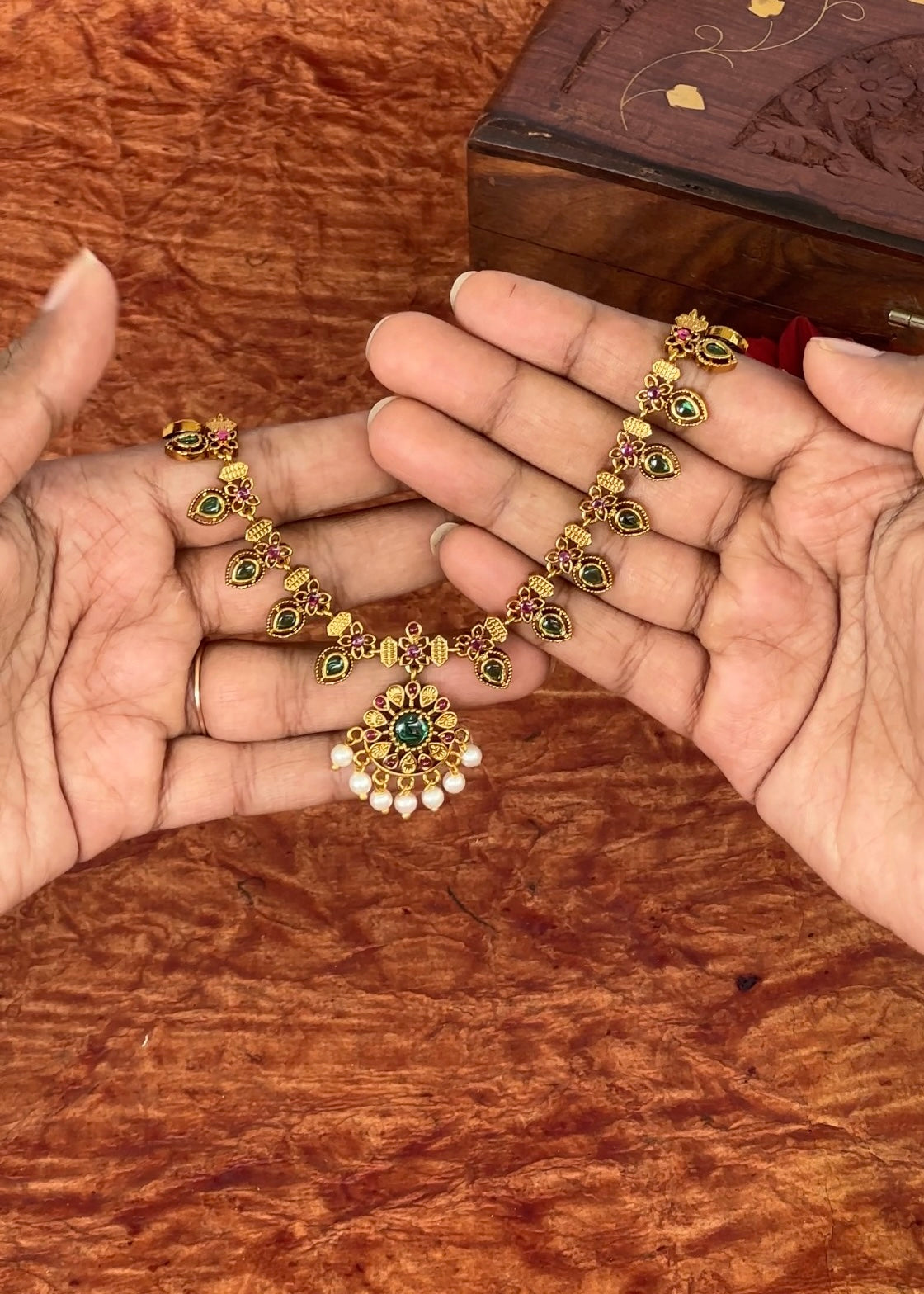 Gold palakka mala necklace with green gemstones held between two hands on a wooden surface