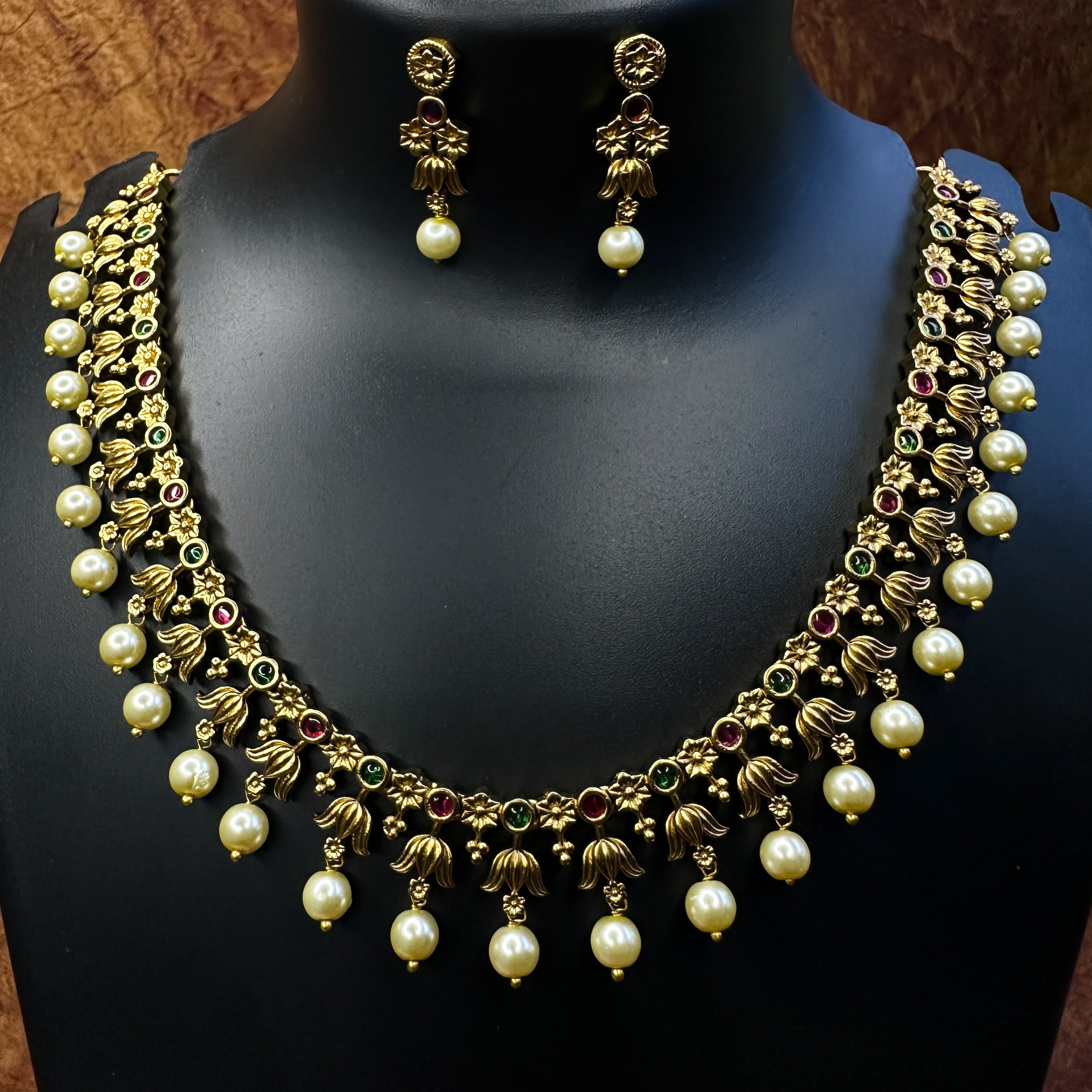 Ethnic Gold necklace with pearl droppings and earrings on a black mannequin with a pink flower and brown background