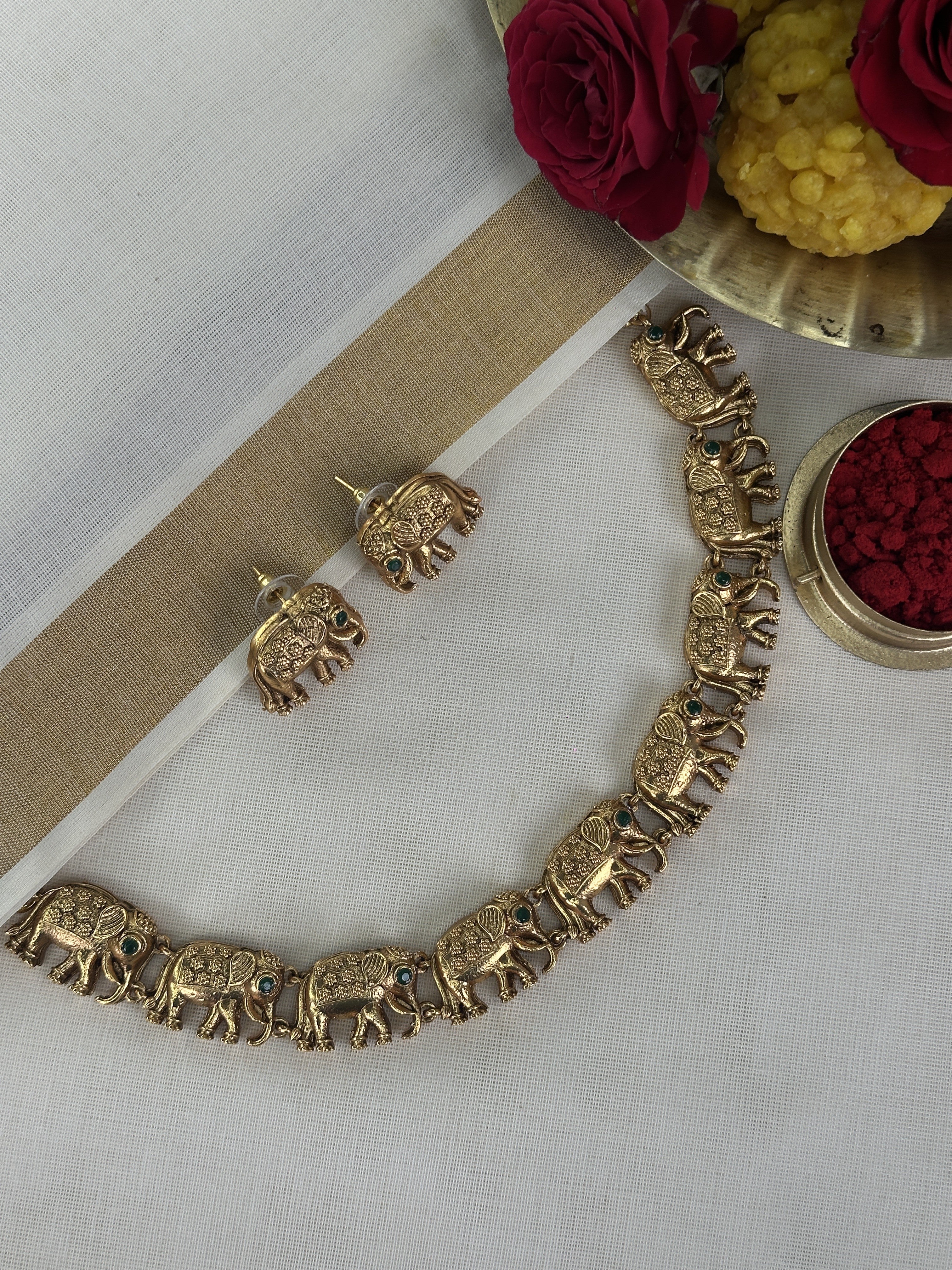 Gold elephant choker with matching earrings displayed on white fabric, surrounded by flowers and a bowl of red flowers