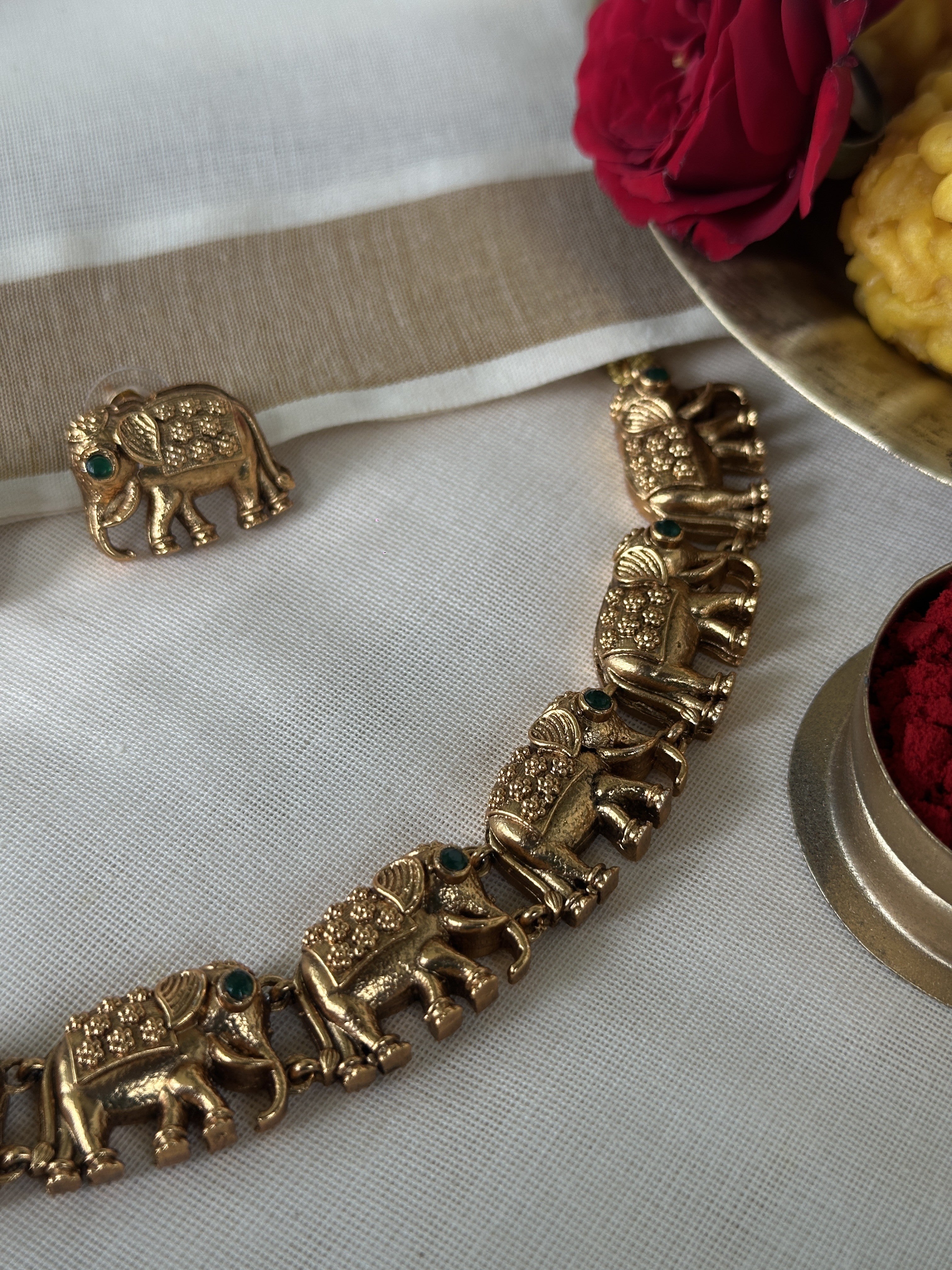 Gold elephant choker with matching earrings displayed on white fabric, surrounded by flowers and a bowl of red flowers